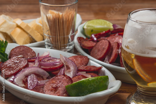 Linguiça frita acebolada com cebola roxa na prato com limão e acompanhada de pão fatiado e cerveja em mesa de madeira no bar ou boteco