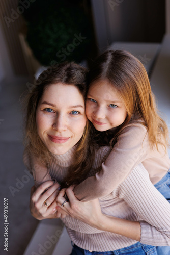 Lovely family mother and daughter embracing and kissing each other indoors. Happy sweet woman with little 6-year-old girl in casual clothers: turtlenecks and jeans sitting on sofa, hugging and smiling