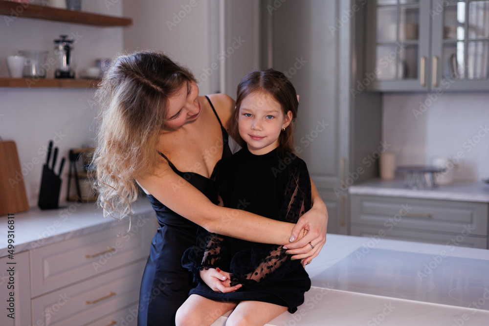 Lovely family mother and daughter embracing and kissing each other in kitchen. Happy sweet woman with little 6-year-old girl in black dresses, family look, hugging and smiling Lovely family mother and daughter embracing and kissing each other in kitchen. Happy sweet woman with little 6-year-old girl in black dresses, family look, hugging and smiling