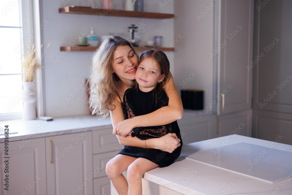 Lovely family mother and daughter embracing and kissing each other in kitchen. Happy sweet woman with little 6-year-old girl in black dresses, family look, hugging and smiling Lovely family mother and daughter embracing and kissing each other in kitchen. Happy sweet woman with little 6-year-old girl in black dresses, family look, hugging and smiling