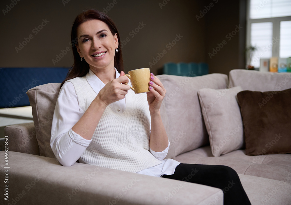 Beautiful relaxed woman sitting on the sofa and enjoying a cup of coffee while picking a comfortable sofa in the furniture store showroom Beautiful relaxed woman sitting on the sofa and enjoying a cup of coffee while picking a comfortable sofa in the furniture store showroom