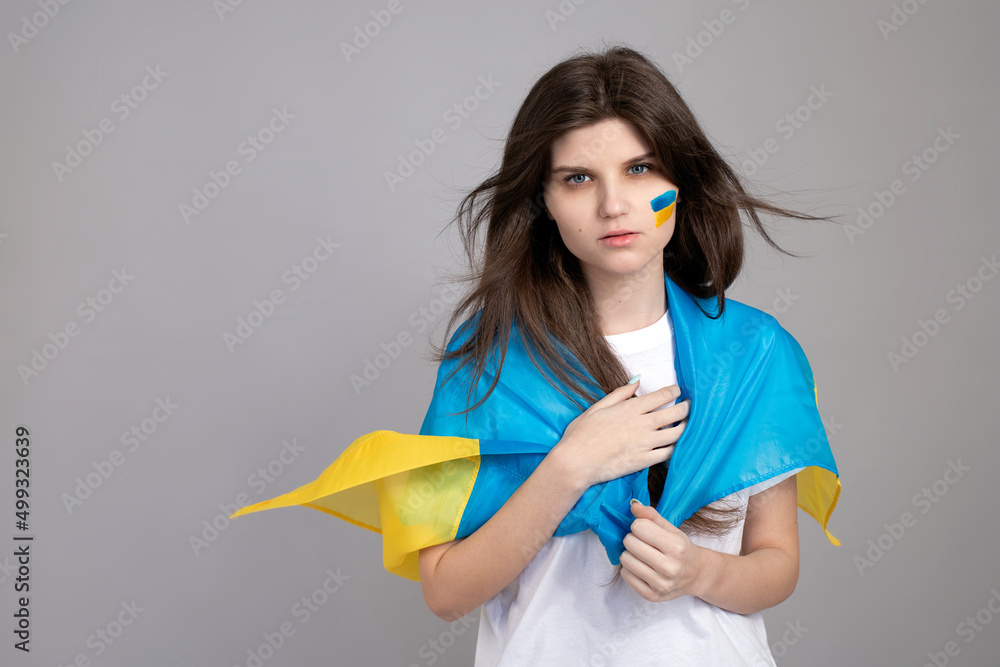 Ukrainian woman stands on a gray isolated background with blue and yellow ukrainian flag on her cheek. the background for your design. Russian invasion of Ukraine, Stop the war. Ukrainian woman stands on a gray isolated background with blue and yellow ukrainian flag on her cheek. the background for your design. Russian invasion of Ukraine, Stop the war.