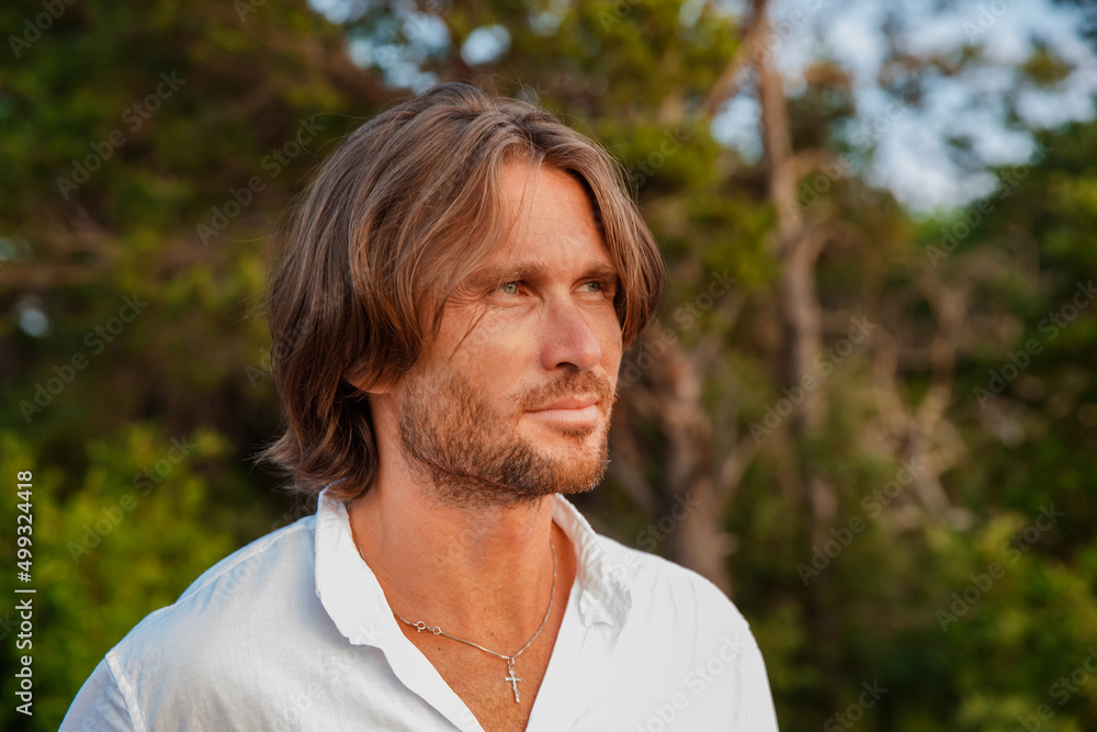 Portrait of handsome brutal man with long hair at sunset, the blue eyes, an easy bristle, he is dressed in a white shirt with a short sleeve, green trees on a background Portrait of handsome brutal man with long hair at sunset, the blue eyes, an easy bristle, he is dressed in a white shirt with a short sleeve, green trees on a background