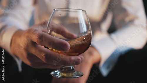 The man sits alone in the room and holds a glass of rum in his hand and shakes glass so that the alcohol flows down the walls of the vessel. Upset man drinking alcohol. Relax with alcohol. Alcoholism