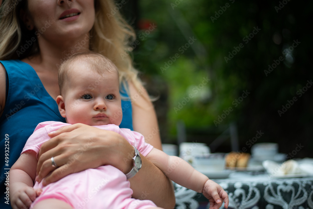 mother holds baby girl with one hand, child looks straight ahead, garden table with porcelain tableware in a blurred background - obrazy, fototapety, plakaty 