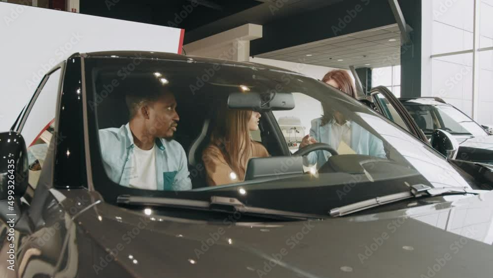 Beautiful multiracial couple sitting inside modern luxury car and examining details of interior. Family choosing vehicle together at auto salon. Saleswoman showing to diverse couple car at showroom. Beautiful multiracial couple sitting inside modern luxury car and examining details of interior. Family choosing vehicle together at auto salon. Saleswoman showing to diverse couple car at showroom.