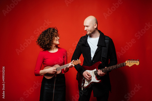 Curly cheerful girl in a red dress plays ukulele. Bald dude playing red guitar.