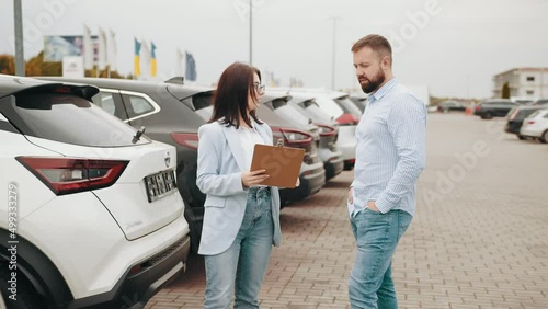 Male customer having conversation with competent saleswoman while choosing car at showroom. Man and woman standing outdoors near row of luxury autos. Female seller helping male client shoosing car. photo