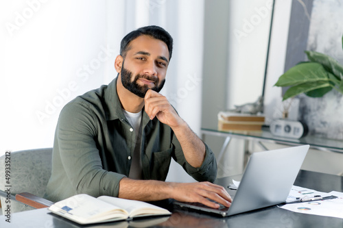 Portrait of a charismatic confident Indian or Arabian man, stylishly dressed, entrepreneur, freelancer or designer, sitting at a desk in a modern office, looking at the camera, smiling friendly