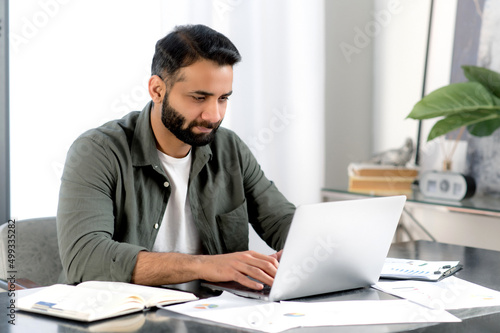 Concentrated confident indian man using laptop in modern office. Successful businessman, freelancer, manager, in casual shirt, browsing email, working and typing on laptop, working on a project