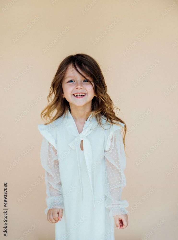 A beautiful little girl in a dress is spinning on a beige background. The concept of a happy childhood, dancing, holidays. Long hair flying, the girl smiles and rejoices.