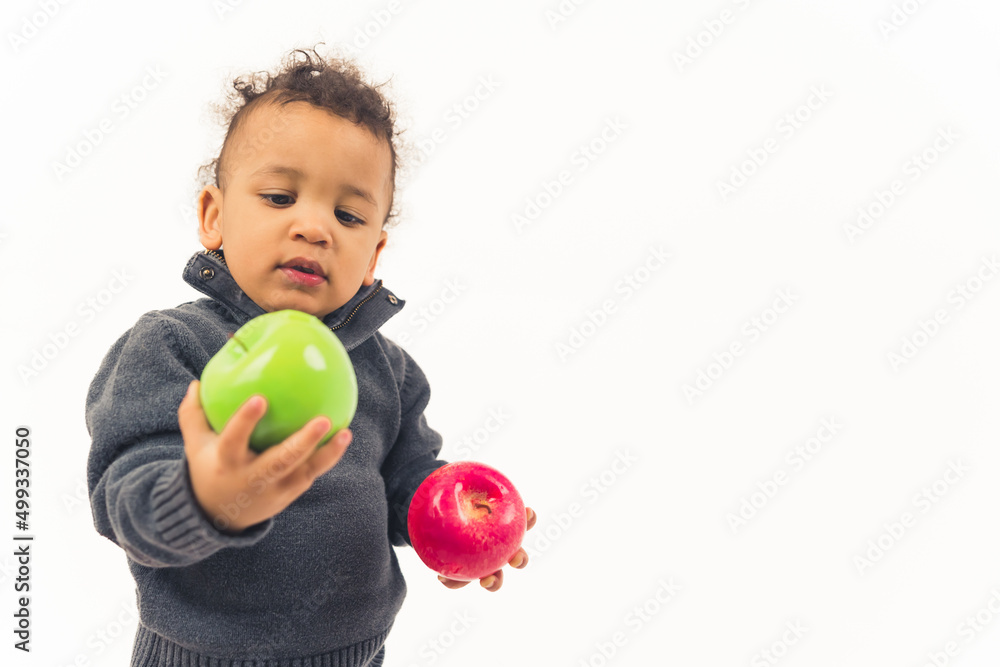Biracial baby boy with curly hair looking at juicy green apple in one hand and holding red apple in the other. Healthy food concept. High quality photo Biracial baby boy with curly hair looking at juicy green apple in one hand and holding red apple in the other. Healthy food concept. High quality photo