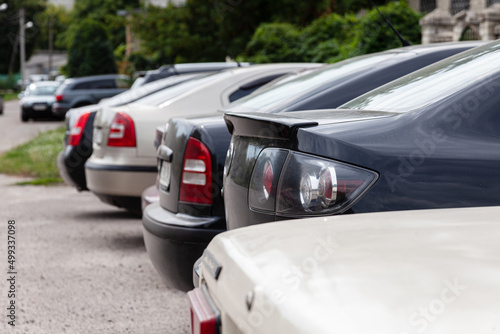 The back of the trunk is visible in the parking space on blurred background. Transportation and parking. Row of cars and vans parked in parking lot during the daytime. © Andrii A