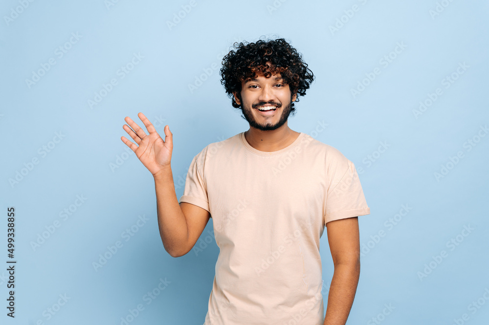 Handsome positive arabian or indian curly-haired guy, wearing casual t-shirt, standing on isolated blue background, looking at the camera, waving hand, welcome sign, smiling friendly Handsome positive arabian or indian curly-haired guy, wearing casual t-shirt, standing on isolated blue background, looking at the camera, waving hand, welcome sign, smiling friendly