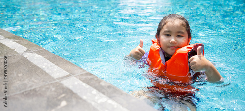 Asian child playing in the pool. Wearing orange life jacket, smiling with thumbs up.