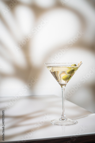 Cocktail vodka martini with green olives on light background with bright beautiful shadows