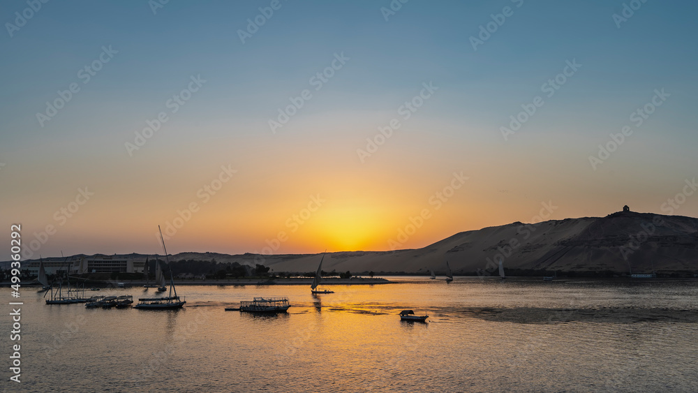Sunset over the Nile. The sun hides behind the sand dunes. The sky is colored orange. Glare on the water. Tourist boats and felucca sail along the river. Egypt. Aswan Sunset over the Nile. The sun hides behind the sand dunes. The sky is colored orange. Glare on the water. Tourist boats and felucca sail along the river. Egypt. Aswan
