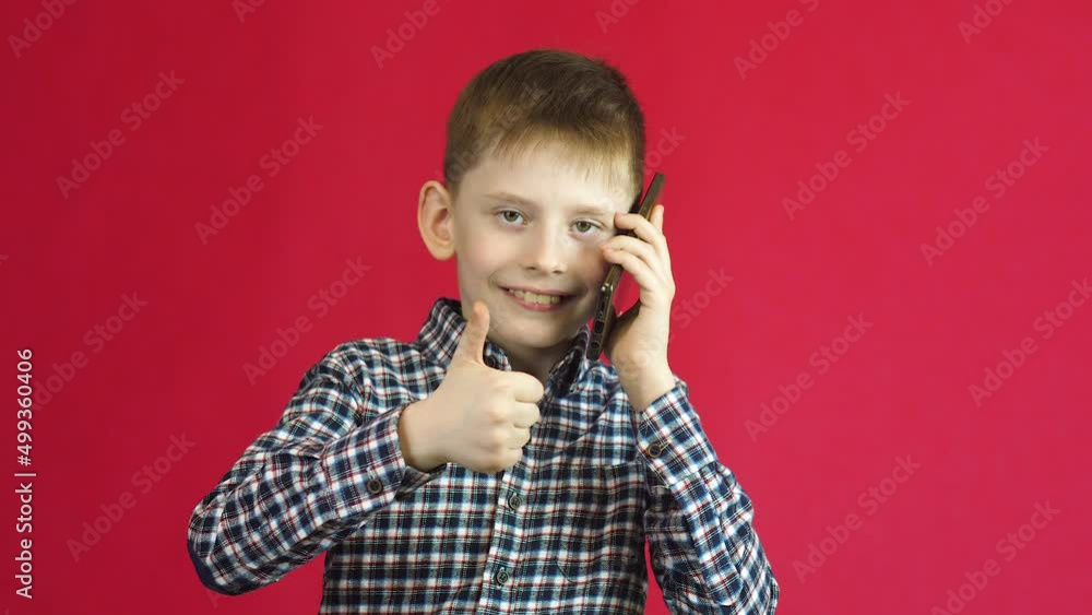 Portrait of an attractive caucasian boy 7-8 years old talking on the phone and showing thumb up smiling. Studio shooting on a red background.Cheerful Child talking on a mobile phone Portrait of an attractive caucasian boy 7-8 years old talking on the phone and showing thumb up smiling. Studio shooting on a red background.Cheerful Child talking on a mobile phone