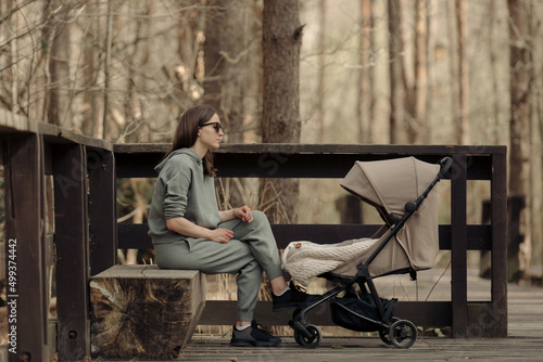 Obraz na płótnie The young mom is relaxing on the wooden bench while her infant child is sleeping in the baby stroller. The mother with her child in the park for fresh air. w salonie