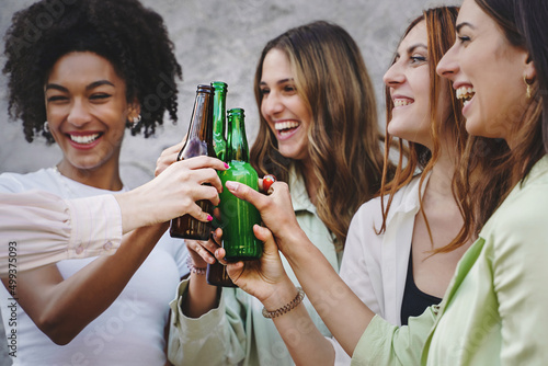 Four young multiethnic girlfriends making a toast with beer bottles while having fun standing outdoors together on the weekend photo