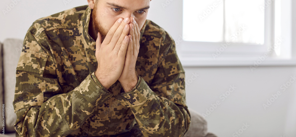 Depressed man recalling war days. Portrait of veteran soldier who has PTSD sitting on couch in military camouflage uniform, holding hands on face and staring at nothing unable to forget horrors of war