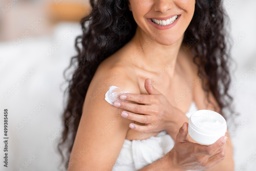 Glad young caucasian brunette lady with long curly hair in towel applies cream on shoulder in bedroom interior Glad young caucasian brunette lady with long curly hair in towel applies cream on shoulder in bedroom interior