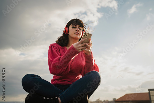Young brunette woman relaxing using mobile smartphone outdoors listening music videoclip from red headphones - tech concept of young people always connected to the internet using modern cellphones.