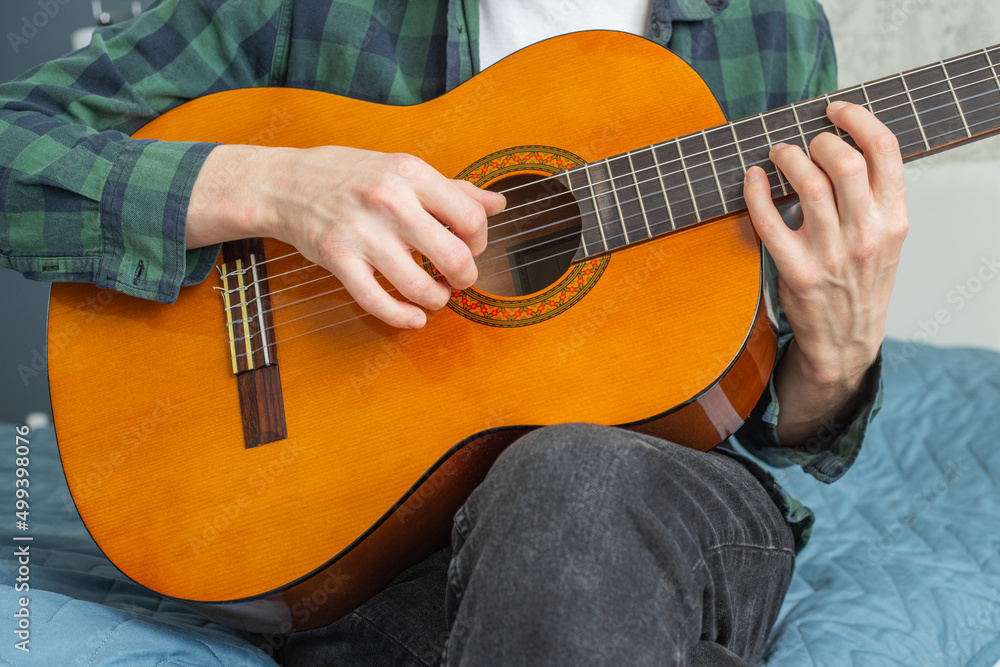 A man in a plaid shirt plays a natural-colored classical guitar sitting on a bed in close-up, selective focus. A male musician plays the guitar. Classical guitar in natural color.