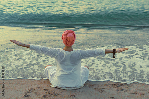 A woman in white clothes and a turban does yoga exercises sitting on the sand by the sea on the morning