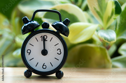 Black alarm clock isolated on blurred nature background. The clock set at 12 o'clock.
