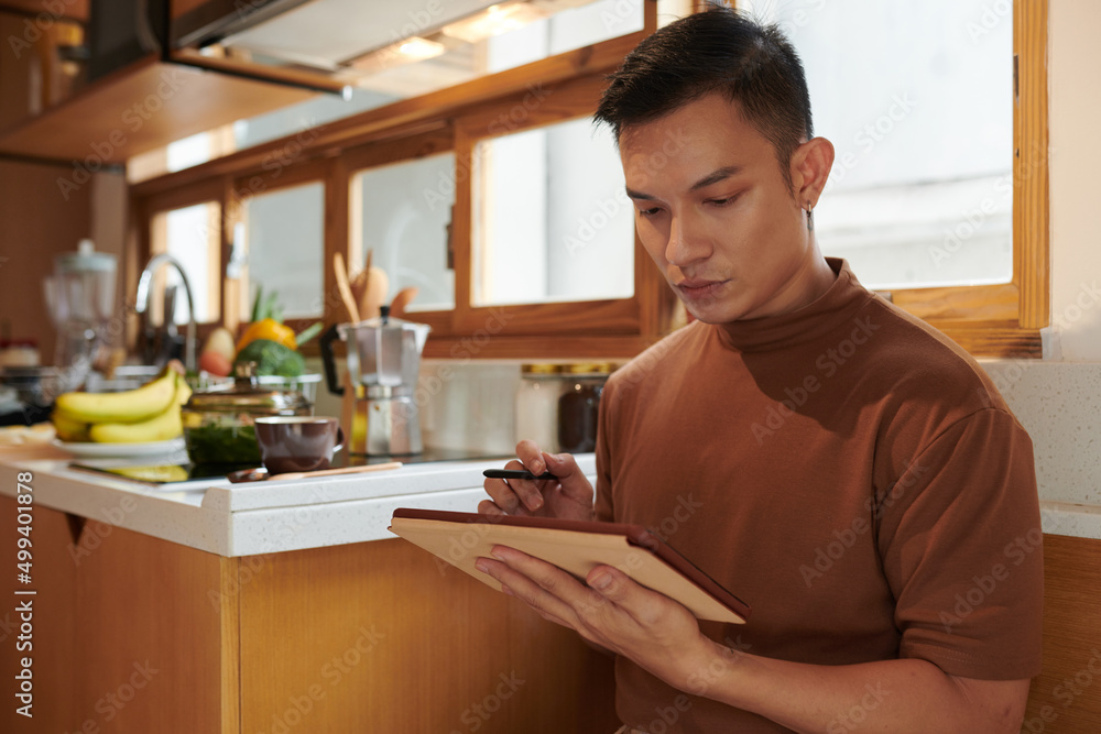 Serious young man standing in kitchen and checking grocery list on tablet computer Serious young man standing in kitchen and checking grocery list on tablet computer