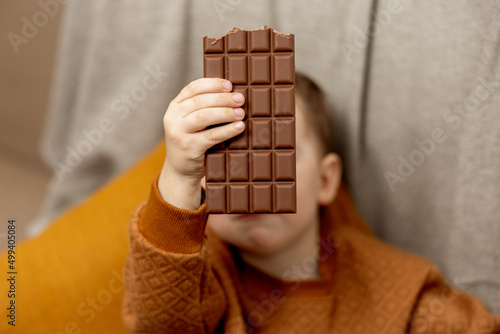 Little adorable boy sitting on the couch at home and eating chocolate bar. Child and sweets, sugar confectionery. Kid enjoy a delicious dessert. Preschool child with casual clothing.