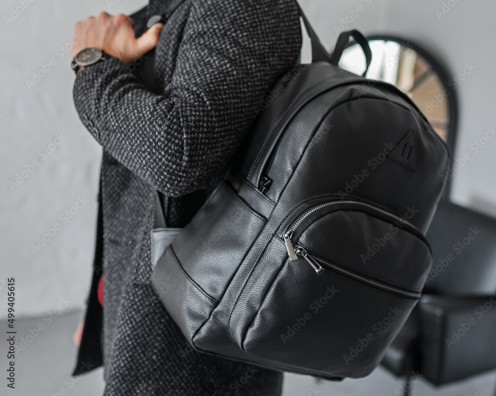 A man holds a black leather backpack on one shoulder. Man s hand