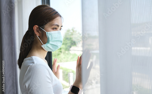 Young woman wearing a medical mask looking through the window. Patient isolated to prevent infection due to epidemic of covid 19, coronavirus. Home isolation concept