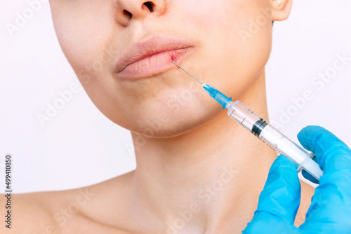 Cropped shot of young woman's face with syringe needle on her lips held by doctor's hand in a blue glove isolated on a white background. Injection of filler in lips. Lip augmentation, enhancement