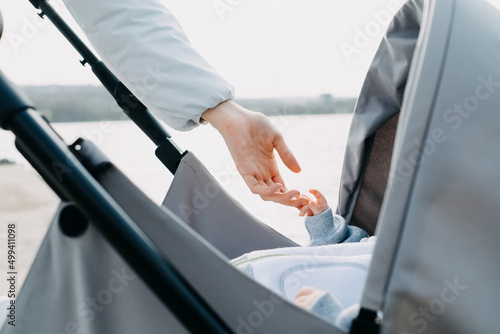 Closeup of baby holding his mother by the hand, lying in a stroller, outdoors.