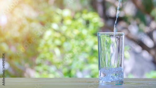The pure water from jug into glass on wooden table on nature background.