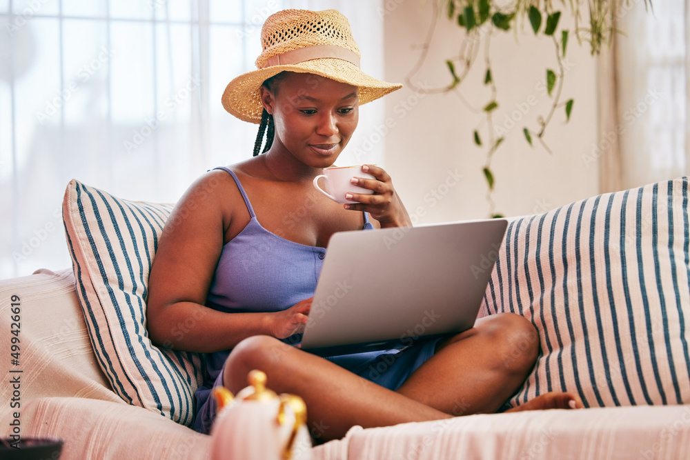 Theres no place like home. Shot of a young woman wearing a sunhat and using her laptop while having a coffee break on the sofa.