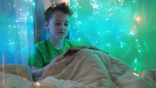 a boy is reading a book sitting on a bed decorated with garlands. multicolored light of lights. a teenager is sitting under a blanket