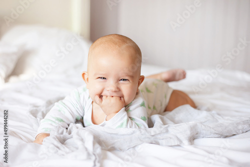 cunning baby lies on tummy on white blanket with fingers in his mouth and smiles