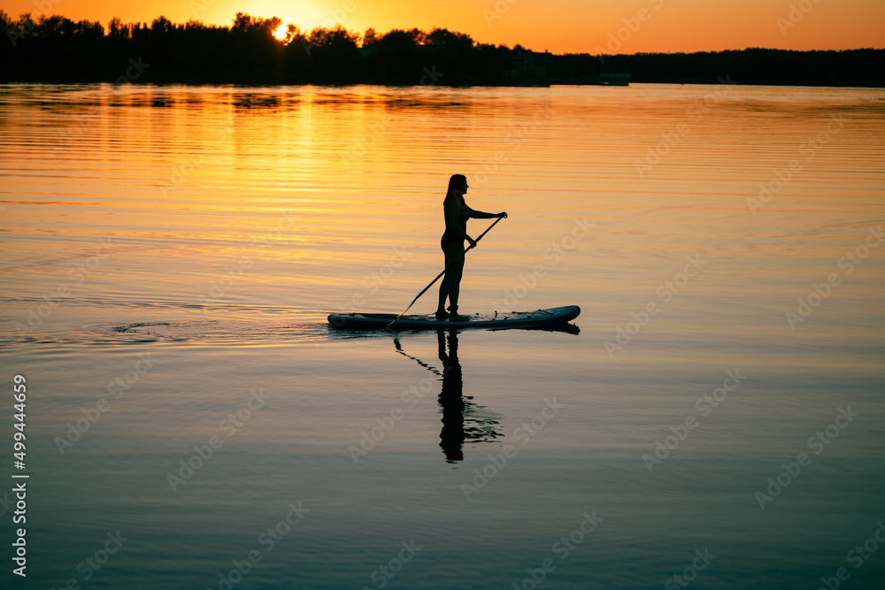 Female sup boarder with oar in hands moving around on river with marvelous sunset in background and reflection on water surface in summer. Active lifestyle.