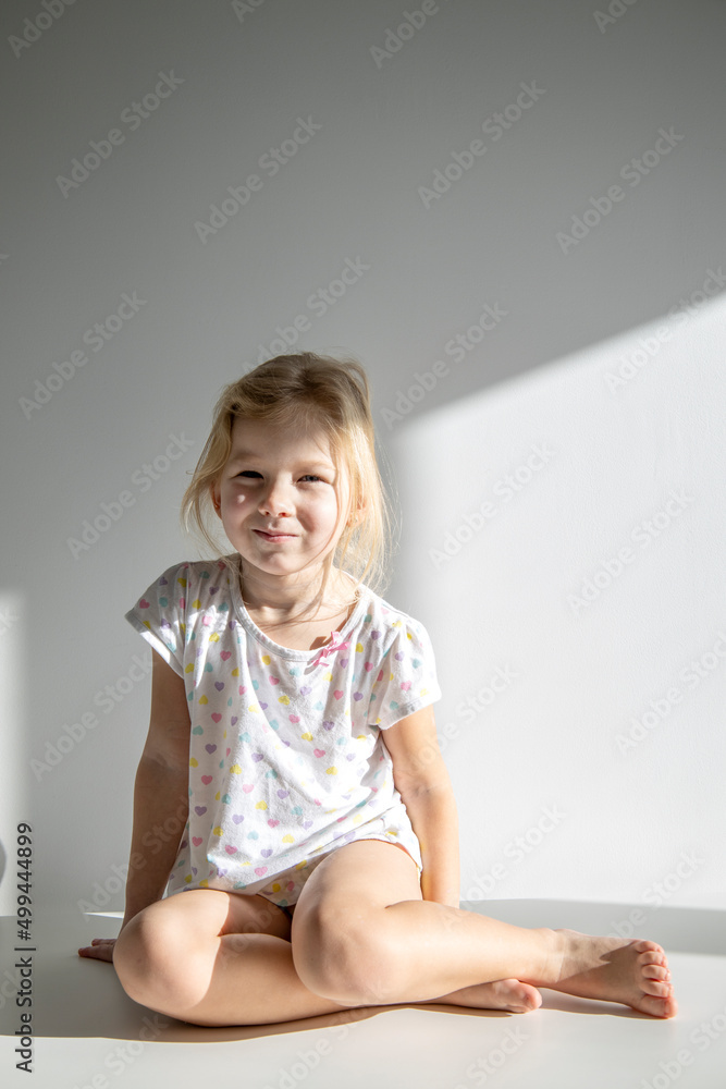 Beautiful child girl blonde sits on a white floor against the background of a white wall.