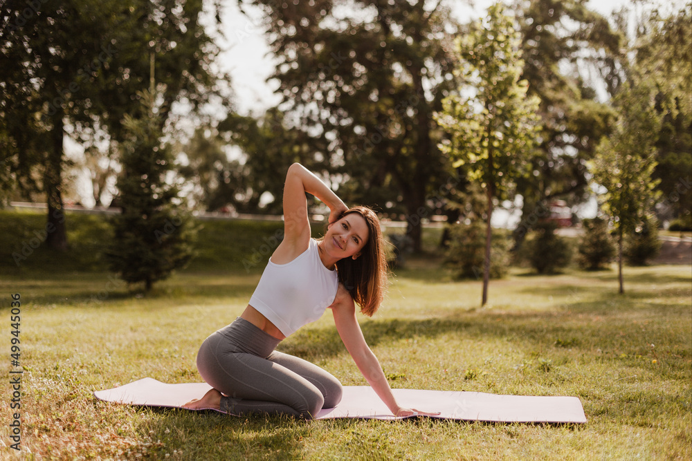Fototapeta premium full length yoga concept. sporty beautiful woman with dark hair sit on her legs with raised bent hand on yoga mat outdoors at park background in sunset. 