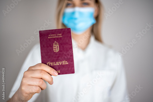 Blond hair nurse in white uniform holding Hungarian passport in front of her. She is wearing blue protective mask on her face. Grey background.
