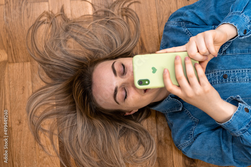 Beautiful young blondie laying on floor and smiling to something on her mobile phone. It has green cover with heart. She is wearing denim shirt. Copy space.
