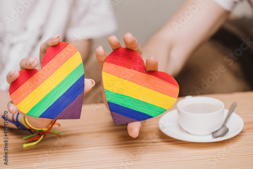 Lesbian couple drinking coffee in a cafe and serfing in smartphone close up. Hands holding rainbow hearts