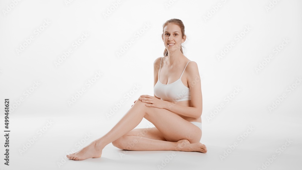 Obraz premium Full shot of slim brunette Caucasian woman in white bikini touches her smooth leg sitting on the floor and looks at the camera smiling on white background | Leg and body care concept