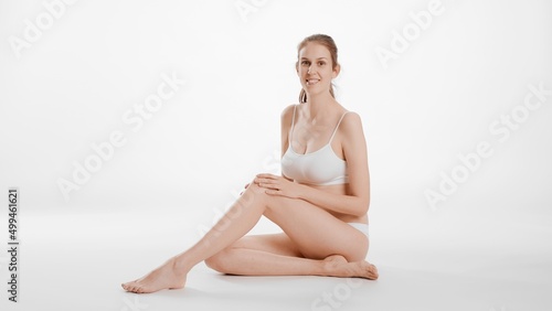 Full shot of slim brunette Caucasian woman in white bikini touches her smooth leg sitting on the floor and looks at the camera smiling on white background | Leg and body care concept