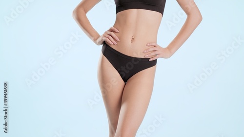 Horizontal medium shot of young slim woman in black bikini who put hands on her hips on blue background | Body care commercial concept
