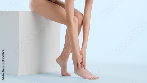 Horizontal medium shot of slim white-skinned woman touches her feets leg sitting on the white podium on pale blue background | Hair removal and leg care concept